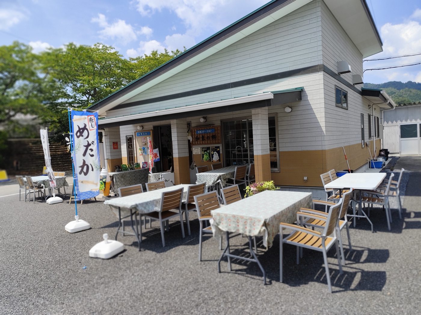 和束の郷の外観。青空の下、軒下と裏の駐車場側に木のテーブルといすのテラス席が並ぶ